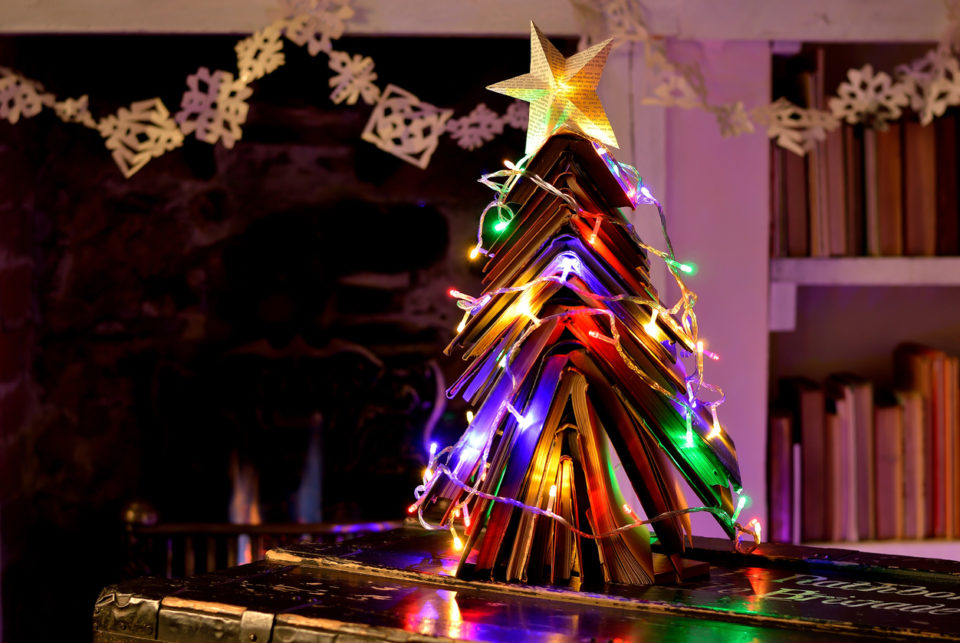 Christmas tree made from vintage books with fairy lights in front of an open flame fire. With star and snowflake chain made from book pages.