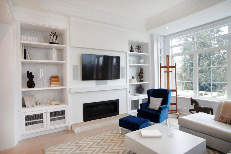 A living room with lots of natural light. There are two built-in bookcases, a fireplace, a wall-mounted TV, a white couch, a white coffee table, a blue chair, and a wooden easel.